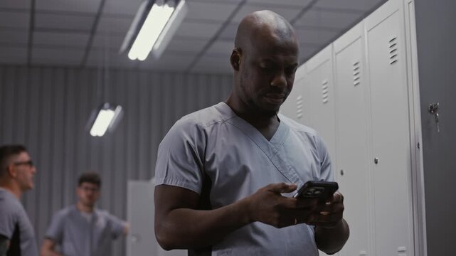 Surgeon in Grey Medical Scrubs Stands by Metal Locker, Checking Messages on Smartphone and Relaxing Before Undressing After Long Workday. Concept of Shift End Routine in Hospital Changing Room.