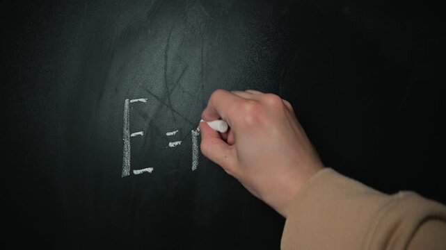 A hand writes the famous equation Emc squared on a blackboard. The action highlights the process of teaching fundamental physics concepts in an educational setting
