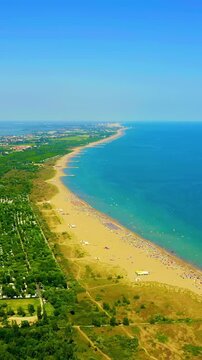Vertical video. Venice, Italy. Punta Sabbioni beach, aerial drone view over golden sand dunes and dense pine forest meeting the turquoise Adriatic Sea under clear summer sky. Drone footage. Rich colo