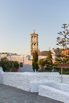 Campanario de iglesia ortodoxa asomando tras muros blancos tradicionales en una isla griega, rodeado de vegetaci&oacute;n bajo un cielo azul.