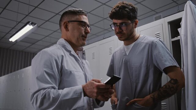 Senior Surgeon Mentors Younger Intern With Tattoos, Using Smartphone to Review Interns Scientific Research Paper and Findings in Staff Changing Area. Medical Education and Mentorship Concept.