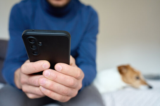 Man using smartphone at home while dog resting on bed in background. Concept of mobile communication browsing home lifestyle and pet companionship
