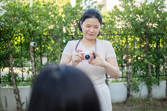 Asian mother taking candid photograph of her daughter outdoors in the garden, documenting precious family moment with her digital camera