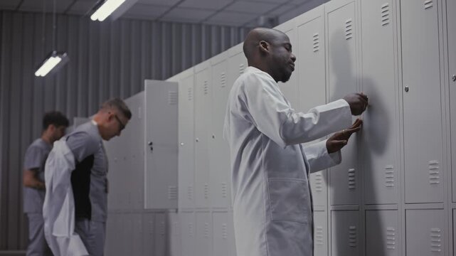Group of Diverse Professional Doctors in Scrubs and White Coats Enter Staff Locker Room, Unlocking Metal Lockers and Starting to Undress After Long Medical Shift. Concept of Healthcare Workflow.