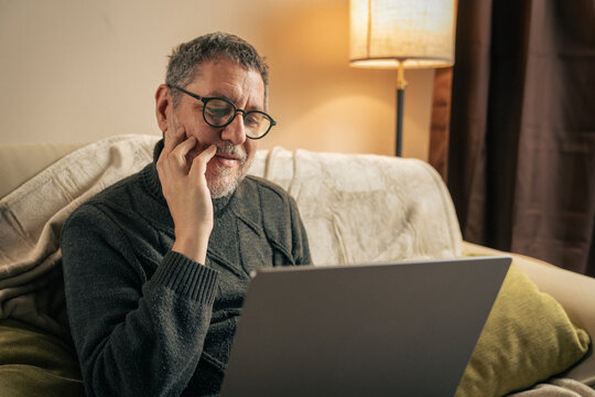 Mature man with glasses sitting on a sofa at home, thoughtfully looking at a laptop screen while researching wellness and health information in a cozy living room.