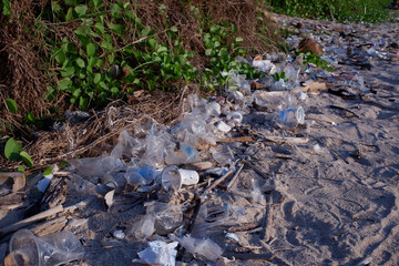 Obraz premium Plastic pollution on sandy beach with green foliage, discarded waste including bottles and cups littering the shoreline