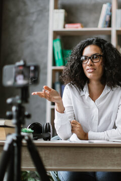 A woman with curly hair and glasses is recording a video with a camera on a tripod in her home office setup