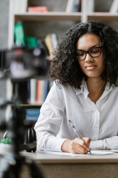 A young woman with curly hair and glasses is writing notes while a camera records her for an online class