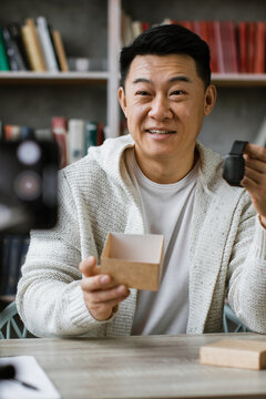 A smiling Asian man is recording a video review of a new smartwatch, holding the device and its open box for the camera