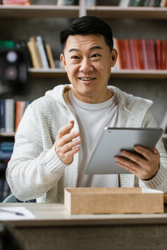 A man is recording himself with a camera while holding a tablet and talking, with a laptop and books in the background