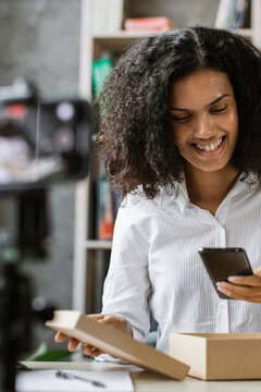 A smiling woman records herself opening a cardboard box while holding a smartphone, with bookshelves in the background
