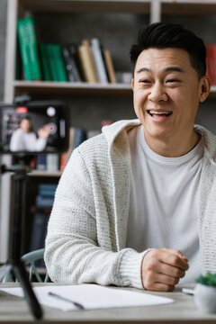 A smiling man waves hello while recording a video on his phone, with a laptop and books on his desk
