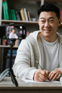 An Asian man records a video blog or online class using his smartphone and laptop in a home office setting
