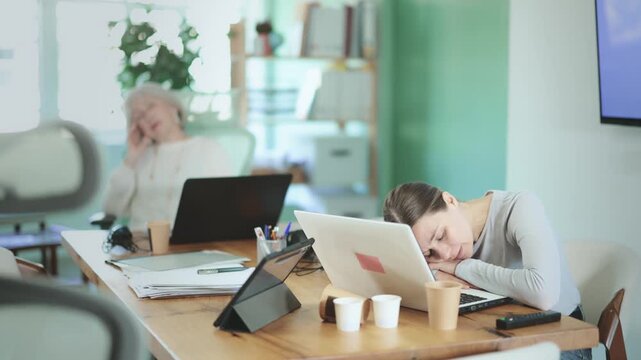 Tired elderly and adult women colleagues falling asleep while working with tablet and laptop in office