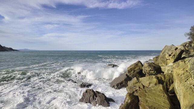 Big powerful ocean waves crashing on a rocky shore in Getaria, Basque Country. Energetic white foam from the sea splashing on the rocks under a beautiful blue sky with some wispy clouds