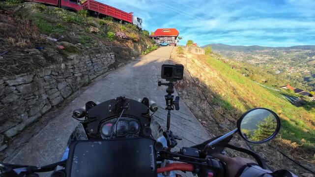Motorcycle ride along narrow mountain road, featuring mounted action camera and dashboard in foreground. Scenic rural landscape with hills, red-roof house, and truck ahead, adventure travel