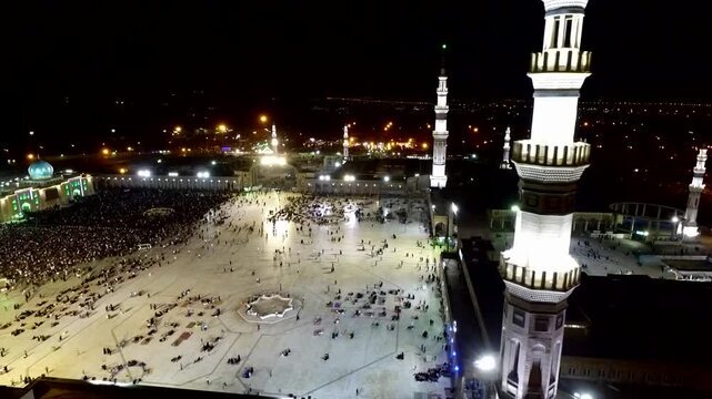  Aerial view highlighting a mosque's tall minarets and illuminated domes, illustrating the fervent devotion of a large number of pilgrims gathered for prayer at the sacred site. 