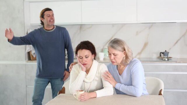 Man is actively quarreling with his thinking wife in kitchen, standing behind her, and elderly mother hugs and calms her daughter 