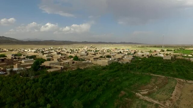  Aerial shot of a rural village nested in a green mountain valley, surrounded by arid hills and fields. A scenic view of human settlement integrated into a rugged mountain landscape in Iran. 