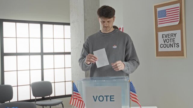 Woman inserting ballot into transparent ballot box on a table inside a voting building while wearing yellow sweater; civic duty solemn.