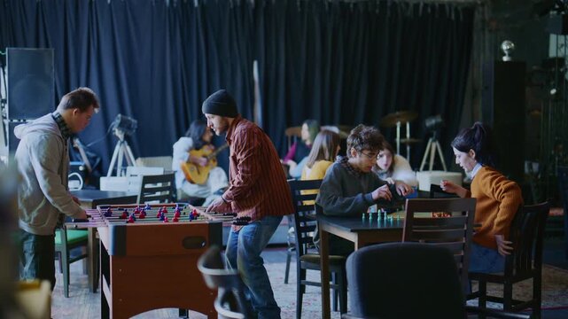 A diverse group of young adults socializes and plays games in a community space. Students enjoy foosball, board games, and music in a relaxed setting.