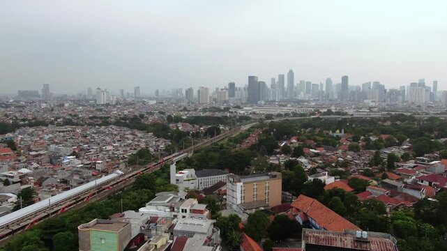 Aerial view of commuter train passing through dense residential housing area with city skyscraper skyline in the background. Concept of urban development, public transportation, and overpopulation