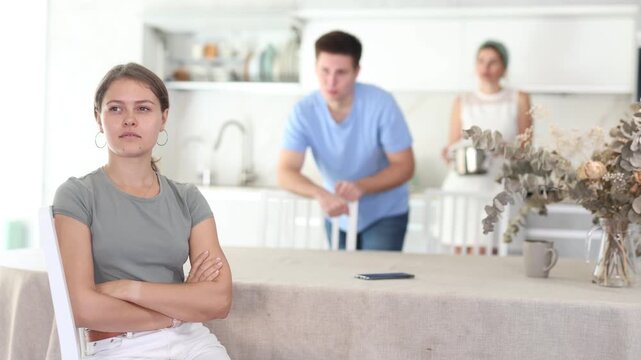 Sad offended young woman sitting at kitchen table during argument with dissatisfied husband and mother-in-law standing behind on blurred background. Concept of family fights 