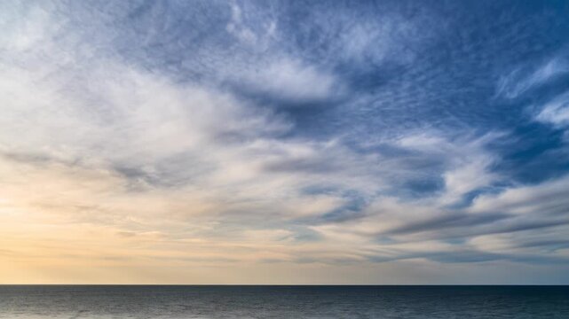 Time-lapse of white clouds moving quickly across a blue sky over the ocean. Dynamic cloudscape above the sea horizon at sunrise. Nature and weather background