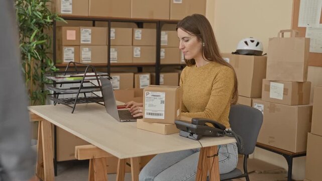 Woman receives package at building desk, smiling as man hands parcel across laptop and shelves of boxes visible; friendly customer service.