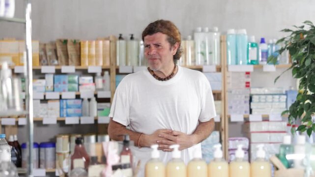 Man in summer clothes is standing in a pharmacy and holding his stomach, he feels cramps and indigestion. Buyer came to a European pharmacy for pills for diarrhea. High quality 4k footage