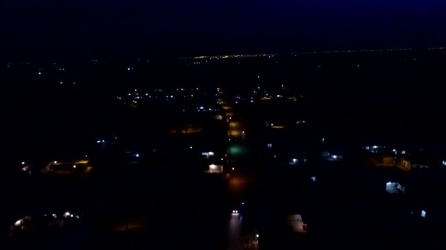  Night aerial view of city lights glowing in darkness. Urban illumination forms patterns across streets and buildings, creating a dramatic nocturnal cityscape scene. 
