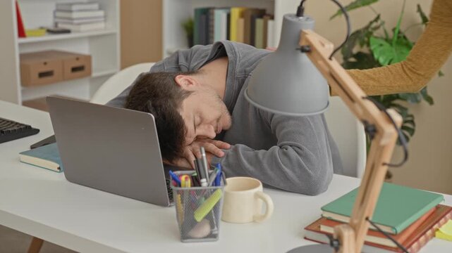 Man at laptop with woman touching his shoulder and leaning over while he glances up from a desk scattered with books and a lamp inside a building office; concern support.