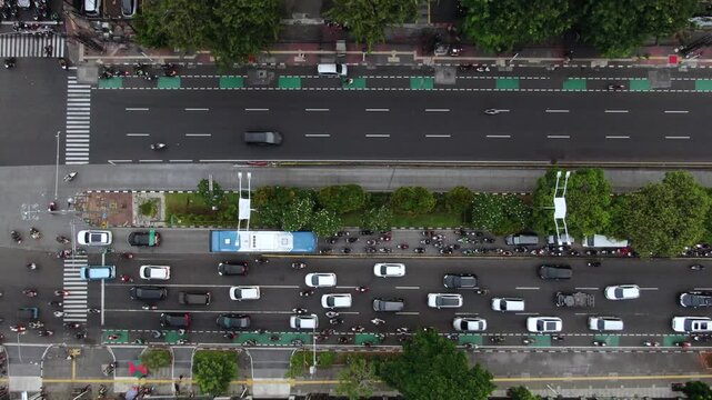 Aerial top down view of busy city main road with traffic flow. High angle shot of urban infrastructure featuring TransJakarta bus, green bike lanes, motorcycle commuters, and metropolitan street