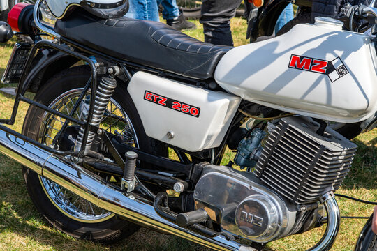 Close-up of a vintage white MZ ETZ 250 motorcycle, showing engine details, chrome exhaust pipe, fuel tank with IFA logo, and black leather seat, shot from a side profile in bright daylight on grass.