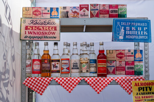 Vintage PRL memorabilia display on a checkered shelf featuring Polish vodka bottles like Wyborowa and Zytnia, cigarette packs including Popularne and Carmen, and retro signs for "Complaints book" and 