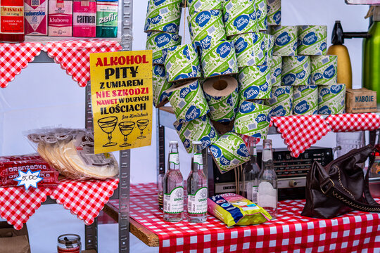 Vintage PRL store display featuring retro cigarette packs, toilet paper rolls on string, Unikat soda bottles, old Diora radio, and a satirical poster on a red checkered tablecloth background.
