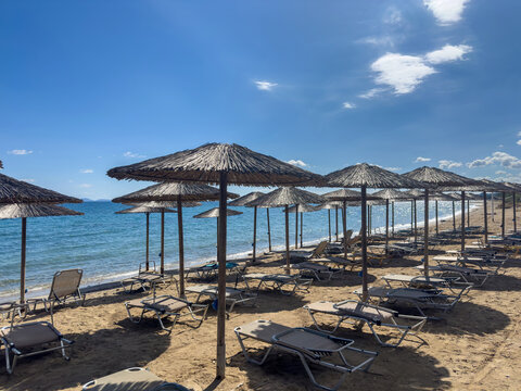 Empty beach loungers under straw umbrellas upon sandy shore within Artemida, Attica, Greece, facing calm Aegean water under bright midday sun and clear blue sky, wide view