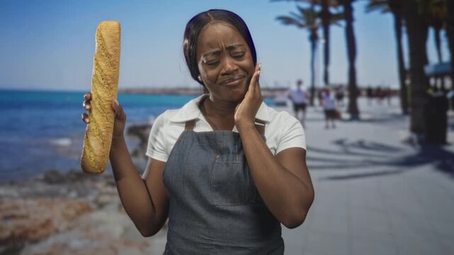 Woman baker in apron holds baguette with left hand and presses cheek with right hand for toothache on street promenade by the sea; discomfort.