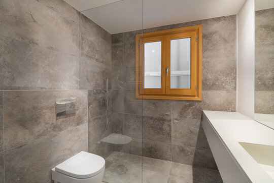 Modern bathroom with gray tiles, a white toilet, and a wooden window. The space is well-lit and features a minimalist design.