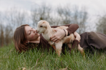 Fototapeta premium woman outdoors grass kiss affection nature countryside field lifestyle moment with goat kid in calm rural setting, tender bond between human and animal, warm light and peaceful atmosphere