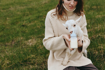 Fototapeta premium Cheerful woman outdoors, feeding a baby goat with a bottle, cozy sweater moment, gentle sunlight, peaceful field, natural colors, wholesome countryside lifestyle, calm connection