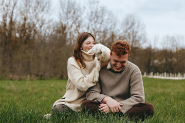 Fototapeta premium Happy couple shares a tender moment with a playful goat kid in an open meadow, sitting on grass, cozy sweaters, warm smiles, peaceful countryside vibe, candid lifestyle scene.