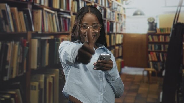 Woman smiling and giving thumbs up while holding smartphone and wearing glasses in a library aisle; confidence study motivation.