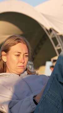 Young blonde woman sitting outdoors during a sunny day showing displeasure and frustration with her facial expressions, puffing her cheeks and looking thoughtful and annoyed