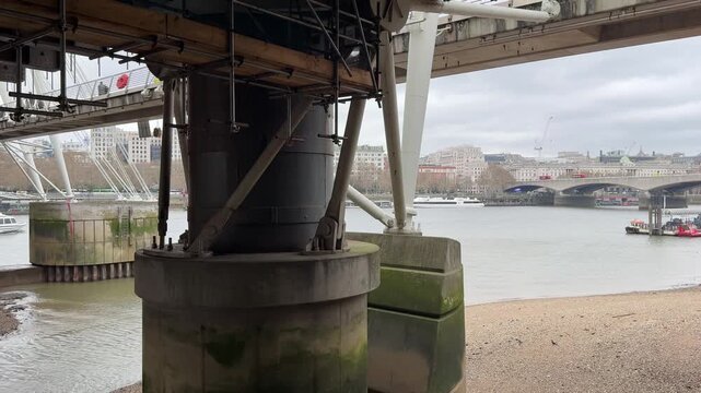 A view from beneath the Golden Jubilee Bridge showing its support structure, the River Thames, and Waterloo Bridge in the distance under an overcast sky