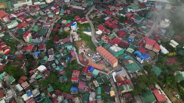 Aerial flyover of a dense hillside neighborhood with colorful rooftops and winding roads across elevated terrain in Baguio City, Benguet, Philippines.