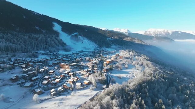 A snow-covered town nestled among sunlit trees with a vast mountain range in the background. Ideal for winter-themed projects or travel content.
