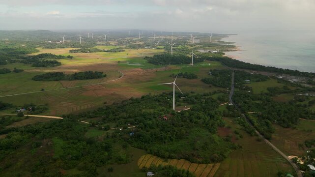 Scenic aerial flyover of multiple wind turbines across coastal farmland with ocean horizon and lush greenery in Guimaras Island, Philippines