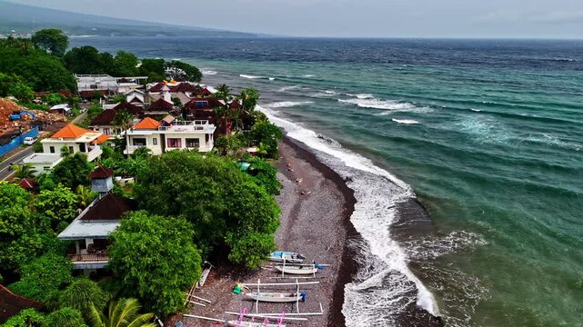 Aerial of Amed's beachfront villas and jukung boats on rocky black sand with choppy grey-green waves. Amed, Bali, Indonesia, Pantai Amed.