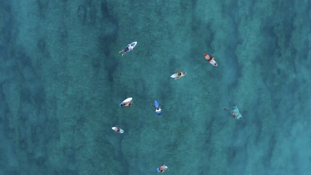 Aerial top-down drone shot over surfers in the lineup as one rides a large breaking wave sending white foam across the reef, capturing both the group and the wave's dynamic energy.